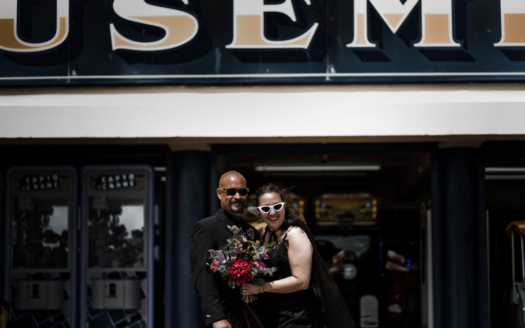 A couple dressed in black pose with a bouquet in front of an amusement arcade.