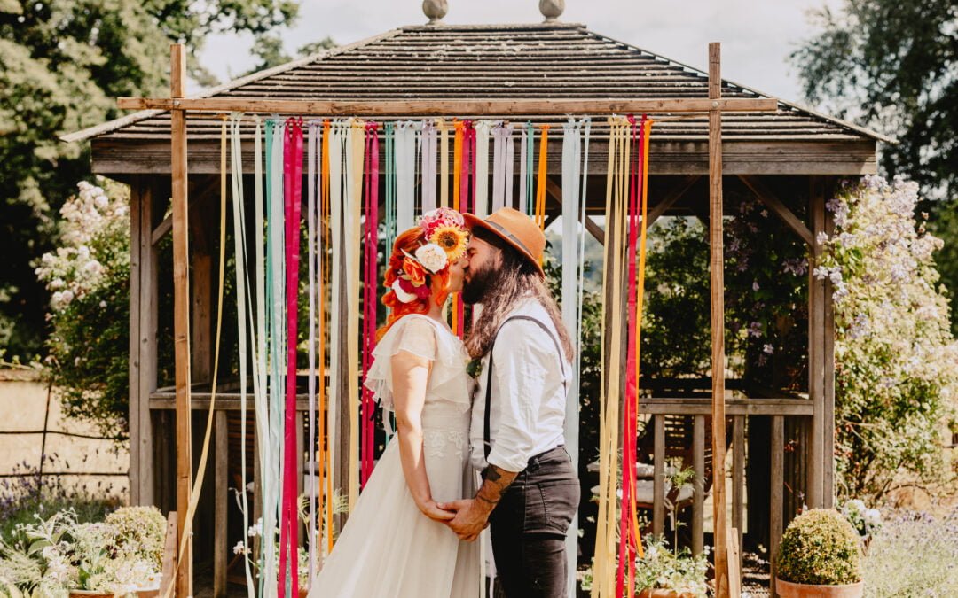 A bride and groom kiss in front of a backdrop of brightly coloured ribbons.